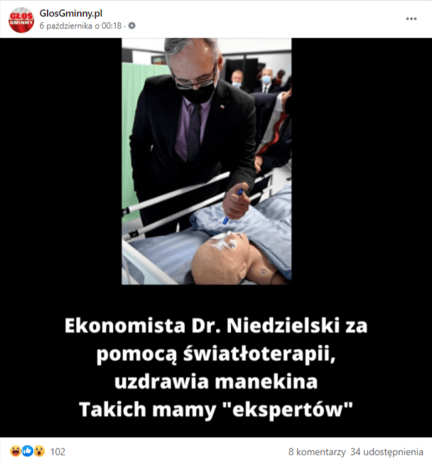 Zrzut ekranu posta na Facebooku. Grafika przedstawia ministra zdrowia Adama Niedzielskiego, który, ubrany w czarny garnitur, fioletową koszulę, ciemnofioletowy krawat i czarną maskę, stoi nad łóżkiem, na którym leży manekin i świeci latarką w jego oko.