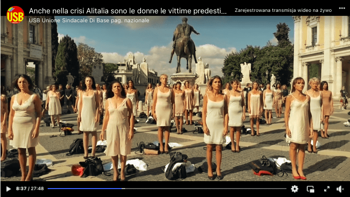 Oryginalny kadr przedstawiający protest włoskich pracownic linii lotniczych. Pejzaż jest taki sam, jak w przypadku wpisu na Facebooku. W kadrze widzimy grupę regularnie ustawionych na miejskim placu kobiet patrzących wprost w kamerę. Każda z nich stoi jedynie w halce, obok zaś mają złożone swoje ubrania. Po bokach kadru widać fragmenty budynków. Z tyłu za protestującymi kobietami widać pomnik na piedestale, przedstawiający postać na koniu. Wokół pomnika ustawione są także inne rzeźby. Niebo jest błękitne z kilkoma chmurami 