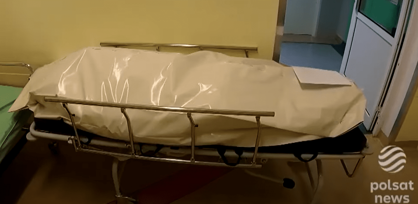 Zrzut ekranu fragmentu materiału wideo, na którym widać biały foliowy worek, w którym znajduje się ciało zmarłej osoby umieszczonej na szpitalnych noszach z metalowym stelażem i kółkami.