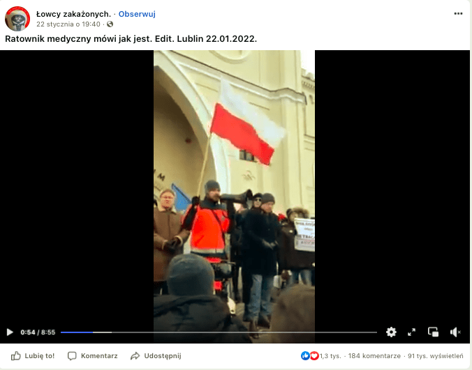 Kadr z wideo przedstawiającego wystąpienie ratownika medycznego na marszu antypandemicznym. W kadrze centralne miejsce zajmuje ratownik ubrany w pomarańczowy kombinezon służbowy, trzymający w jednej ręce megafon, a w drugiej trzonek powiewającej biało-czerwonej flagi. Wokół niego są też inni protestujący