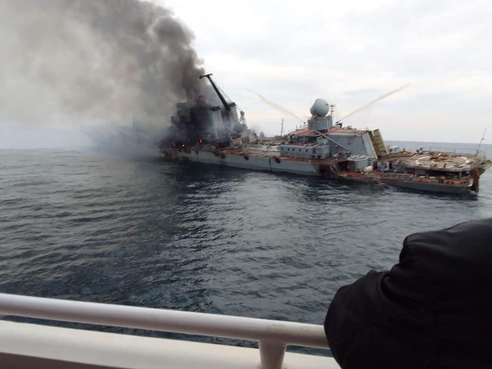 Zdjęcie przedstawiające krążownik Moskwa. Okręt przechylony jest na swoją lewą burtę. Widzimy go od tyłu z lewej strony. Z przodu jednostki unosi się dym.