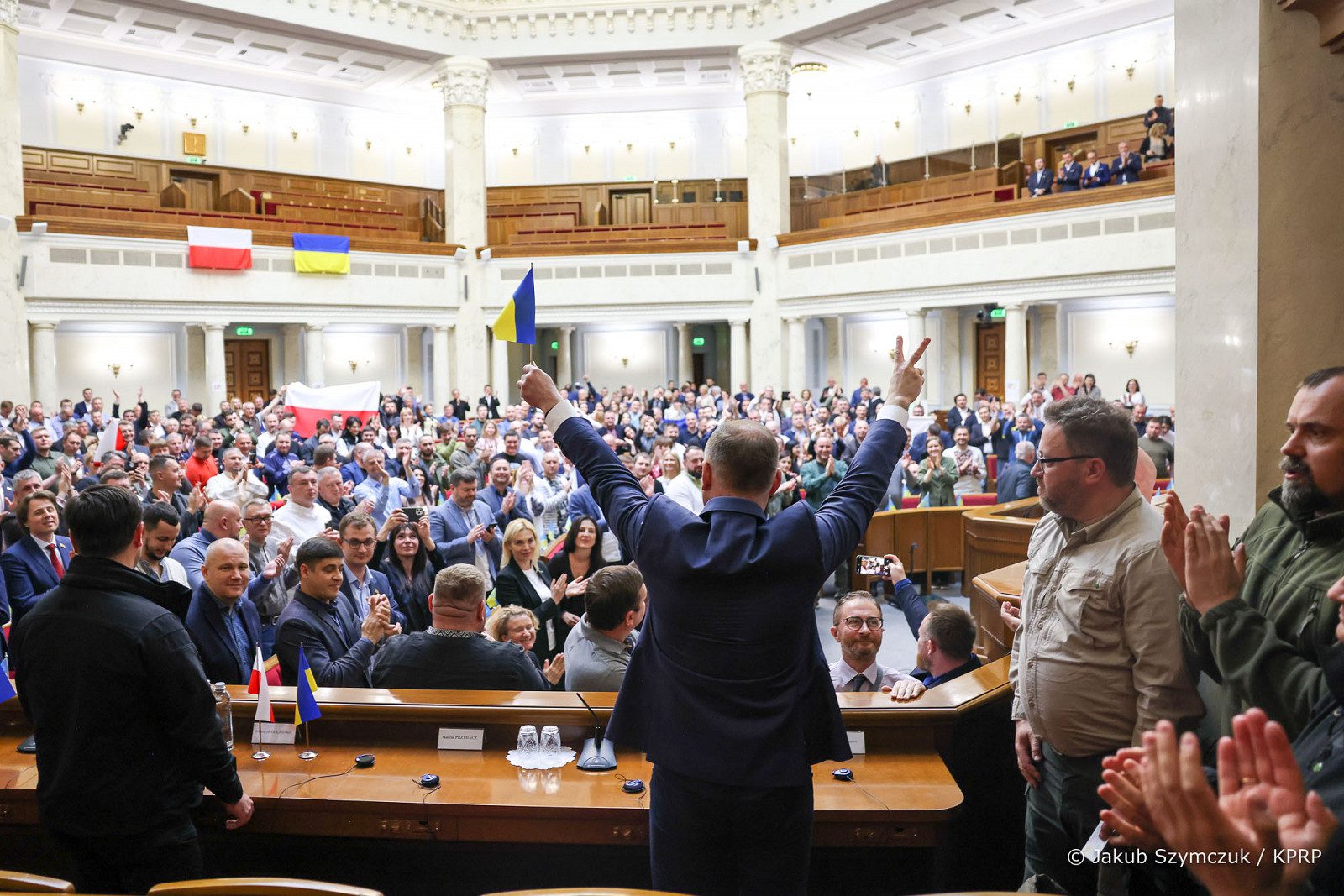 Andrzej Duda przed Radą Najwyższą Ukrainy. Prezydent ubrany w garnitur stoi plecami do zdjęcia. Obie ręce ma wzniesione do góry w geście “V”. W lewej dłoni trzyma małą, ukraińską flagę. Z prawej dłoni ma wyciągnięty palec wskazujący i środkowy w geście „Victoria”. Przed prezydentem widać zgromadzony tłum polityków bijących mu brawo. część zgromadzonych trzyma polskie flagi. Flaga Polski i Ukrainy wisi także na balustradzie piętro wyżej