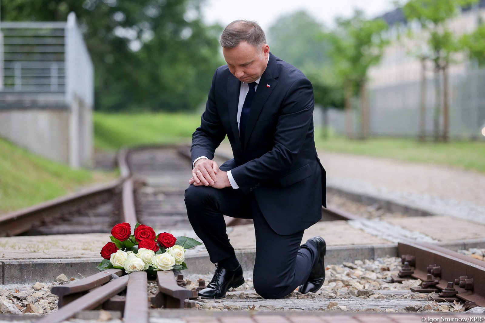 Zdjęcie przedstawiające klęczącego Andrzeja Dudę na torach kolejowych w KL Auschwitz. Prezydent położył kwietny wieniec na jednym z torów.