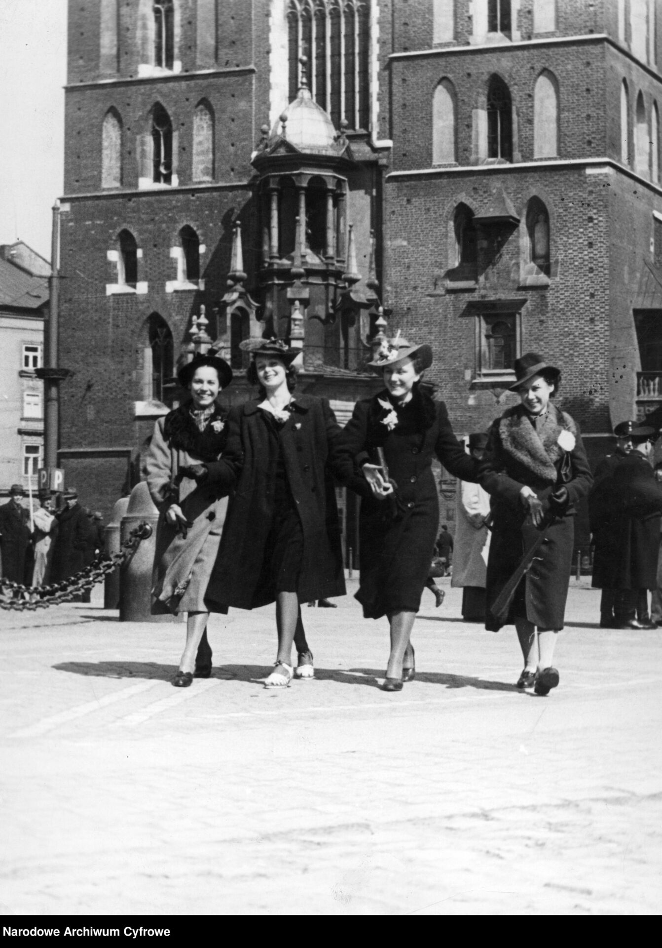 Czarno-białe zdjęcie z 1940 roku, ze zbiorów Narodowego Archiwum Cyfrowego. Widoczne są uśmiechnięte cztery kobiety, spacerujące pod Kościołem Mariackim w Krakowie. 