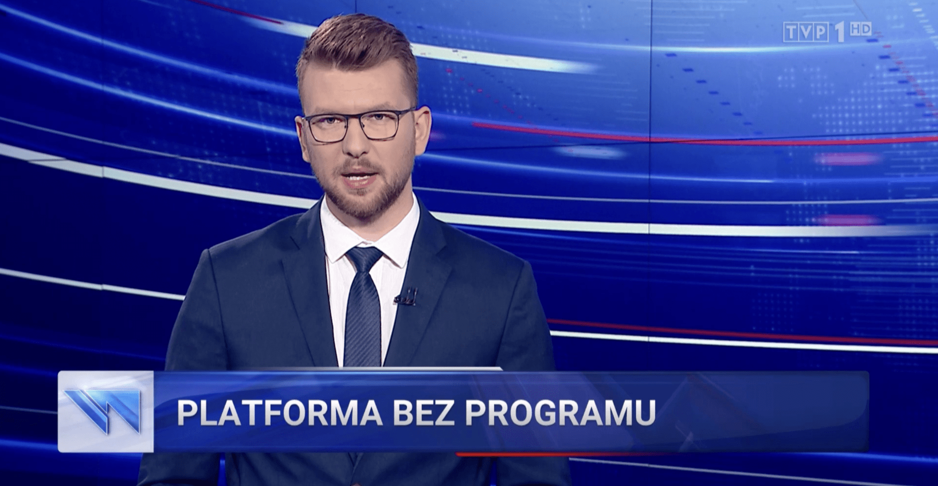 Zrzut ekranu z fragmentu „Wiadomości” TVP1 zamieszczonego na stronie wiadomosci.tvp.pl. Widoczny jest mężczyzna w okularach, w białej koszuli, granatowej marynarce i krawacie. Na dole widnieje tzw. pasek o treści „PLATFORMA BEZ PROGRAMU”.