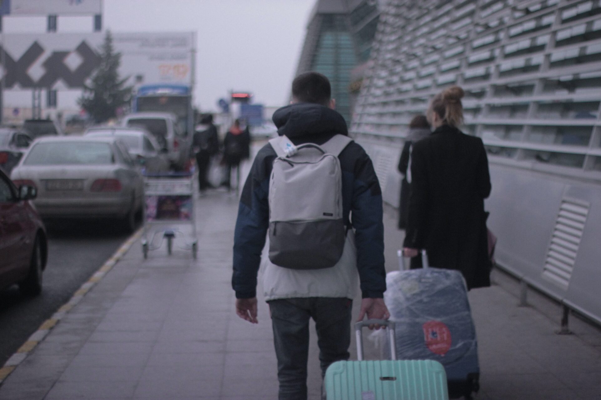 Dwie osoby, mężczyzna z plecakiem w granatowej kurtce i kobieta w czarnym płaszczu, ciągną za sobą walizki, podążając na lotnisko.