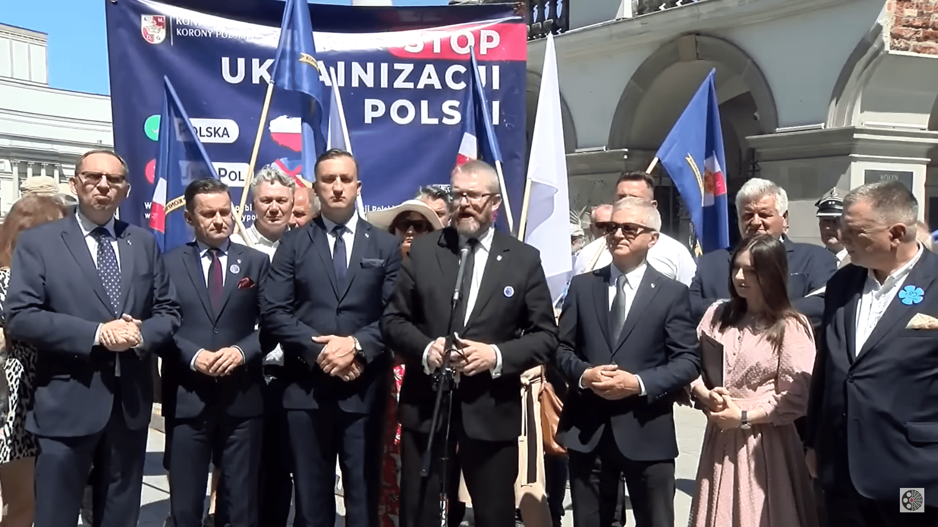 Grzegorz Braun stojący w gronie kilku osób na tle flag, baneru i Grobu Nieznanego Żołnierza w Warszawie
