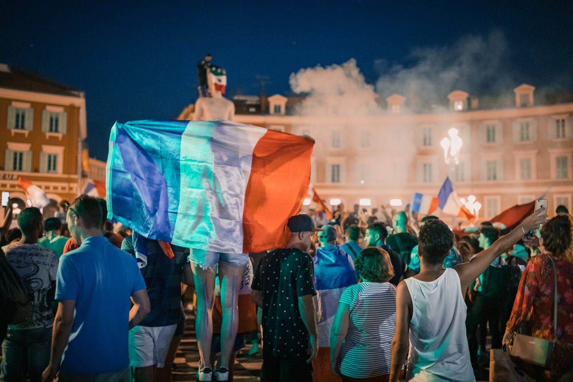 Zgromadzenie ludzi na placu, jedna osoba trzyma za plecami rozwiniętą flagę Francji.