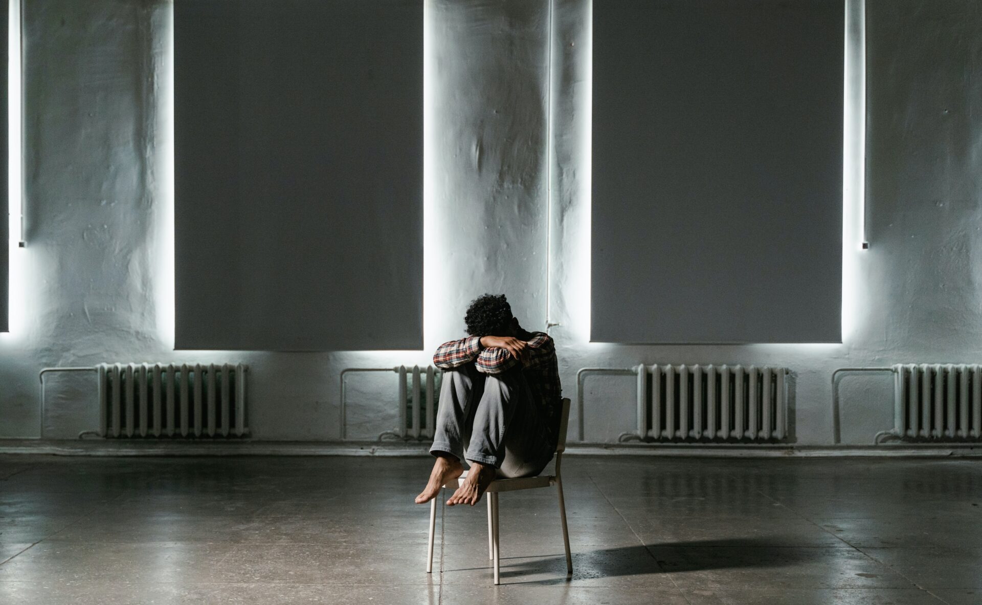Mężczyzna siedzi skulony na krześle w pustym pokoju z zasłoniętymi oknami.