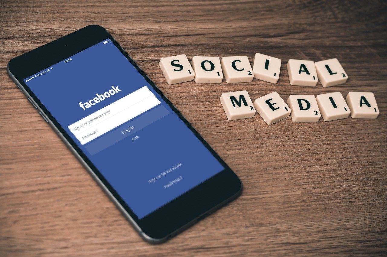 Telefon leżący na stole, na ekranie panel logowania do Facebooka. Po prawej stronie napis “social media” ułożony z literek z gry Scrabble.