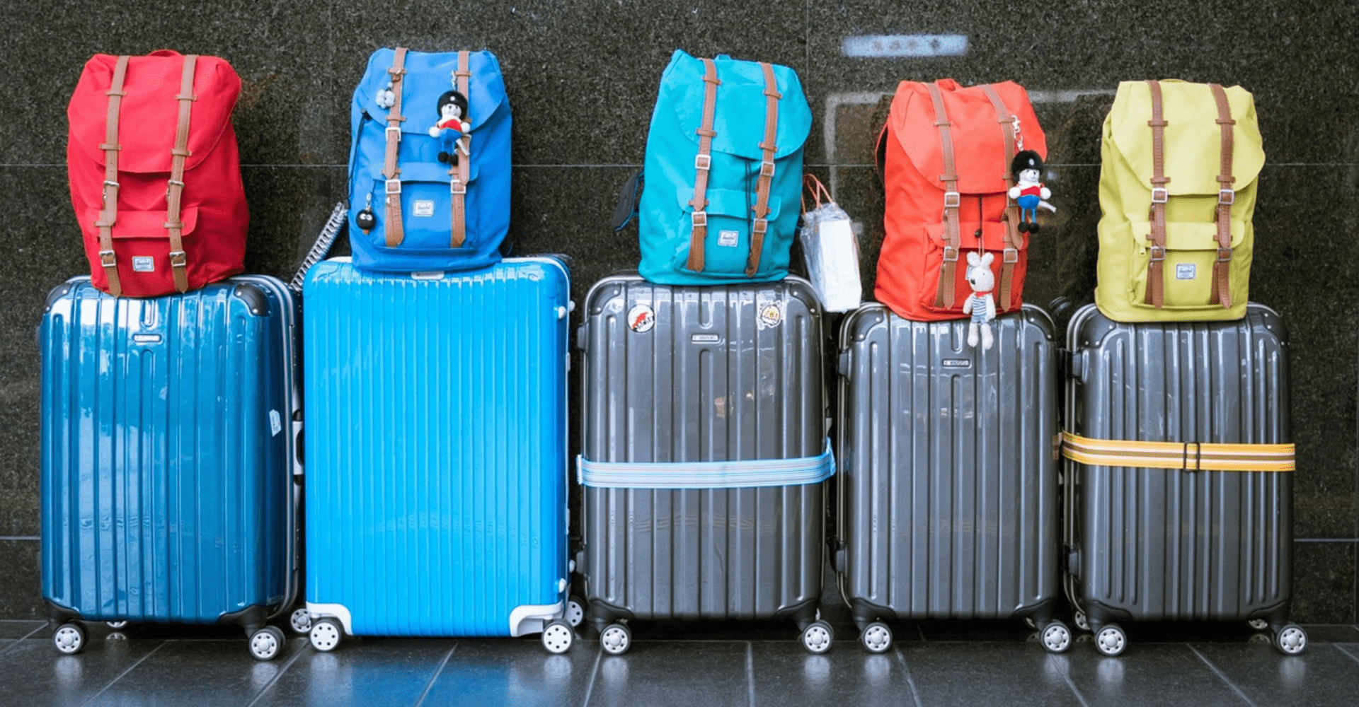 5 dużych walizek, na których położono pięć plecaków w różnych kolorach (czerwonym, niebieskim, turkusowym, pomarańczowym i żółtym)