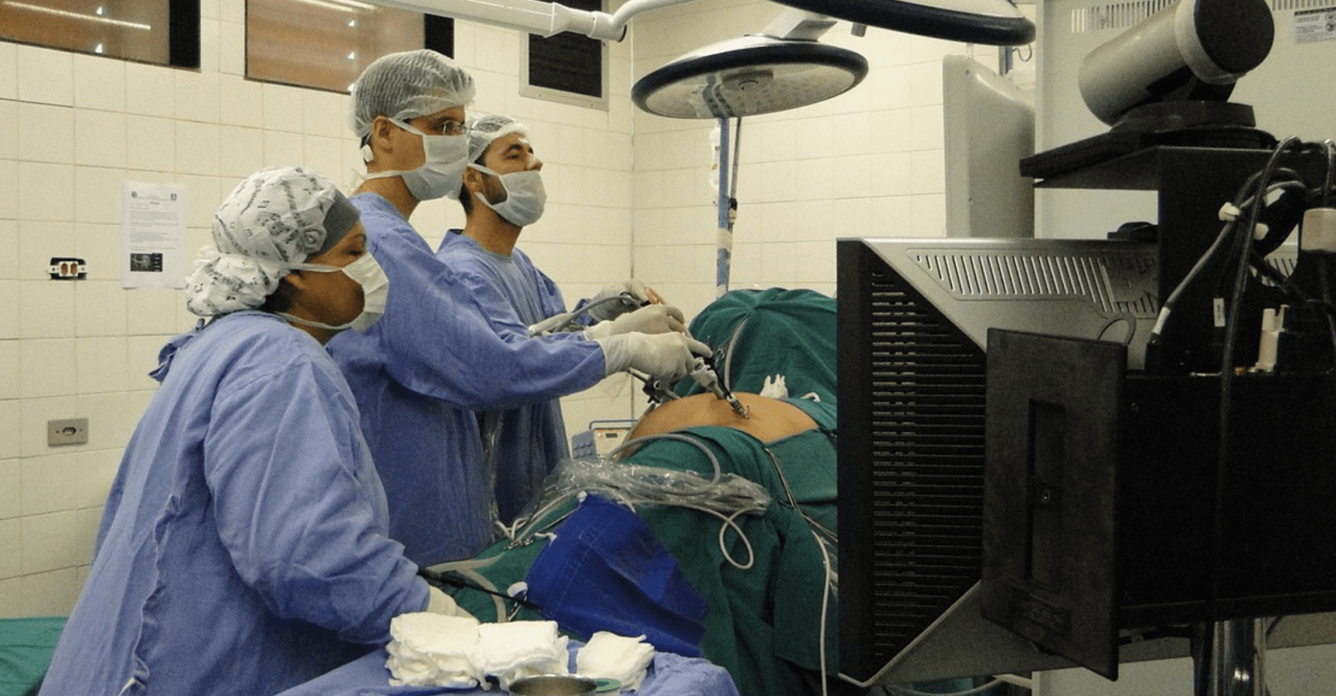 Sala operacyjna. Trzy osoby operują jedną, która leży na stole operacyjnym.