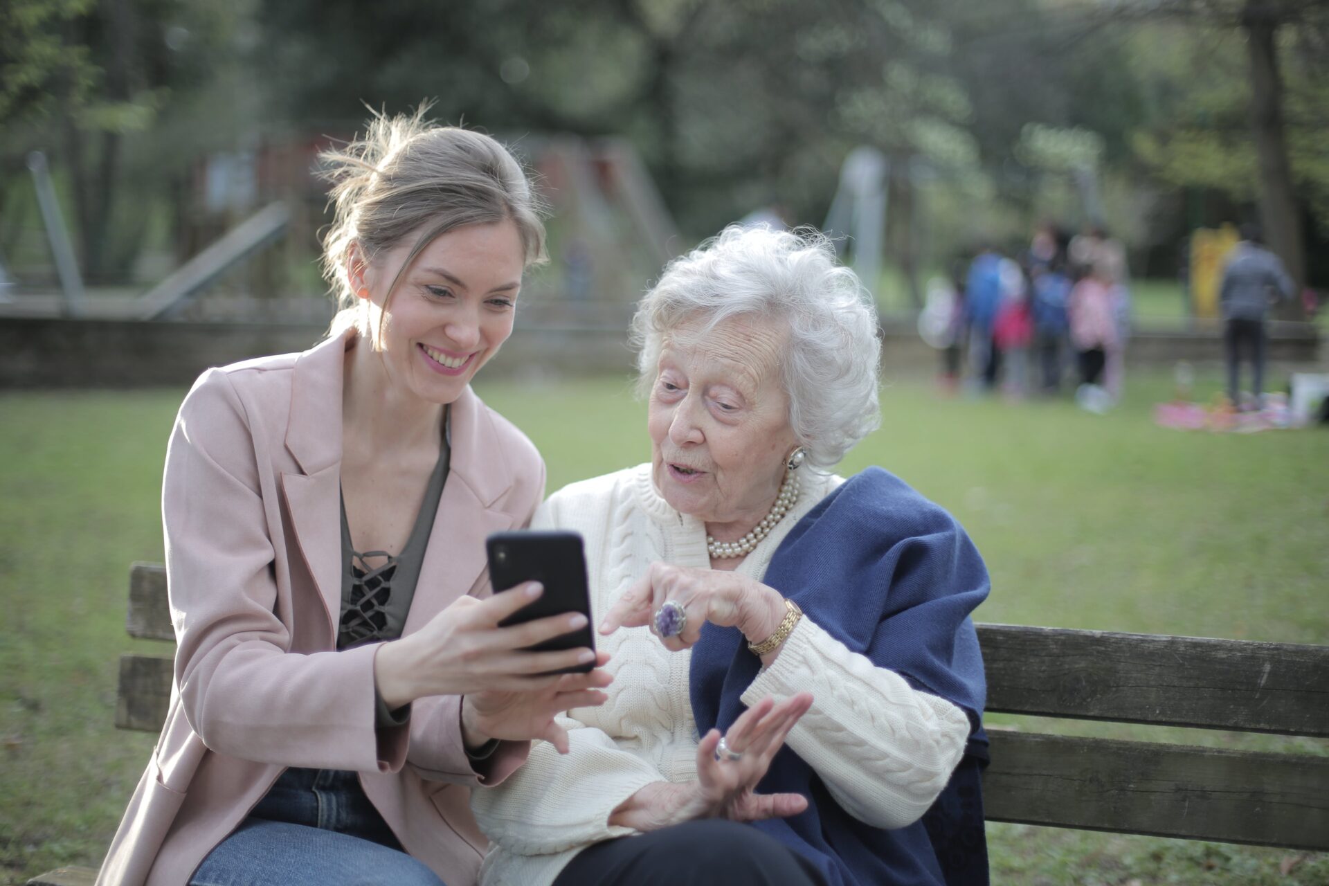 Seniorka siedząca na ławce wskazuje na smartfona trzymanego przez młodszą osobę siedzącą obok