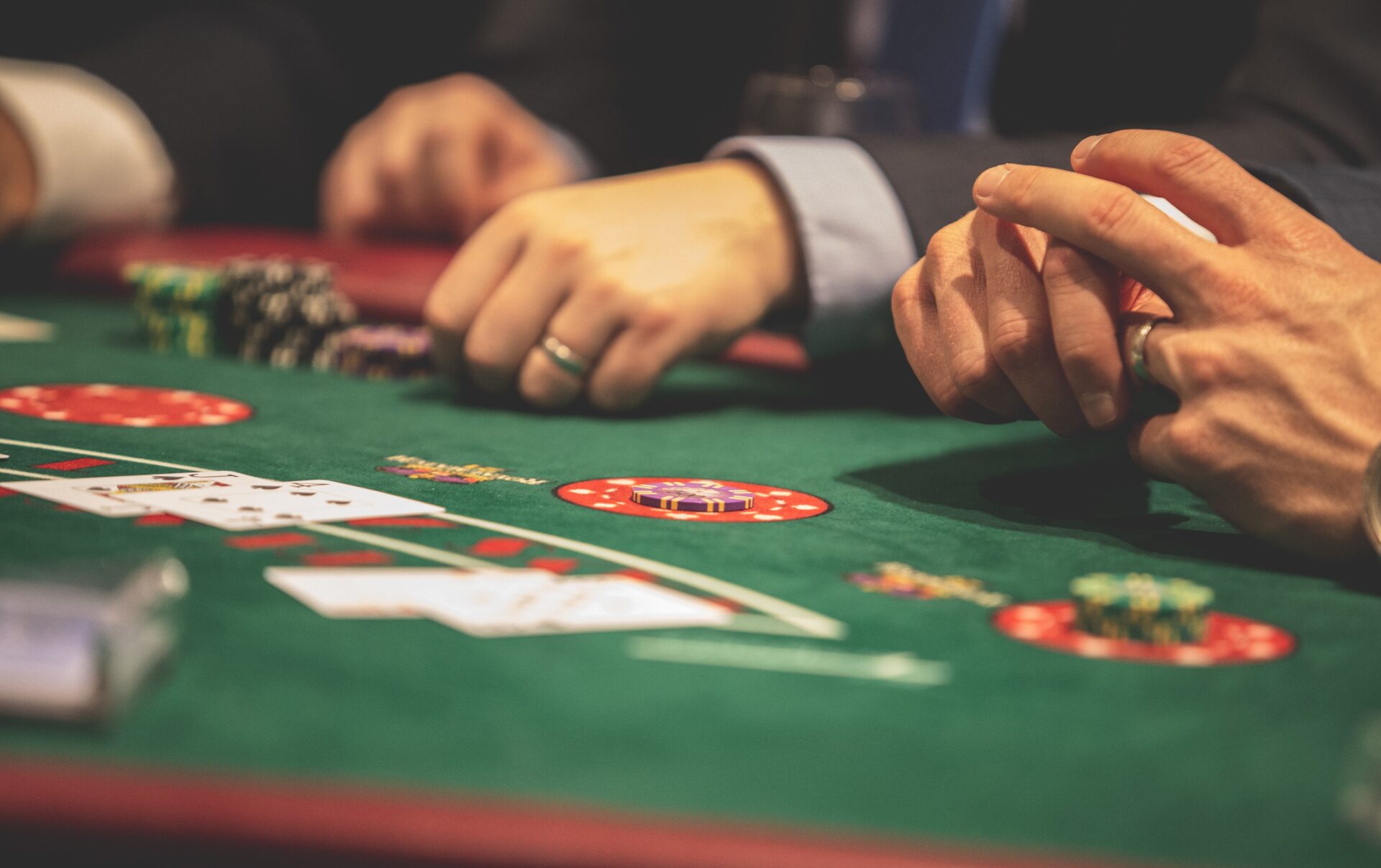 Stół do gry w pokera, na nim karty i dłonie dwóch osób w garniturach.