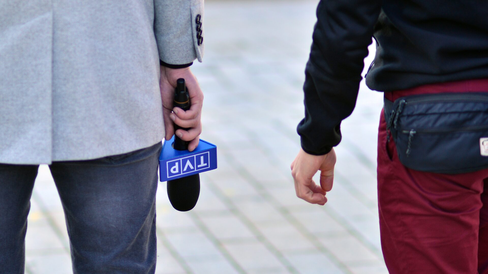 Dwie postaci ukazane od pasa w dół. Idą po chodniku, osoba po lewej stronie trzyma w ręce mikrofon z napisem TVP.
