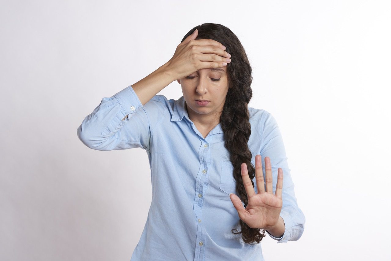 Kobieta w błękitnej koszuli z długim warkoczem po prawej stronie. Prawą ręką łapie się za czoło, z powodu bólu głowy, lewą ma wyciągniętą do przodu.