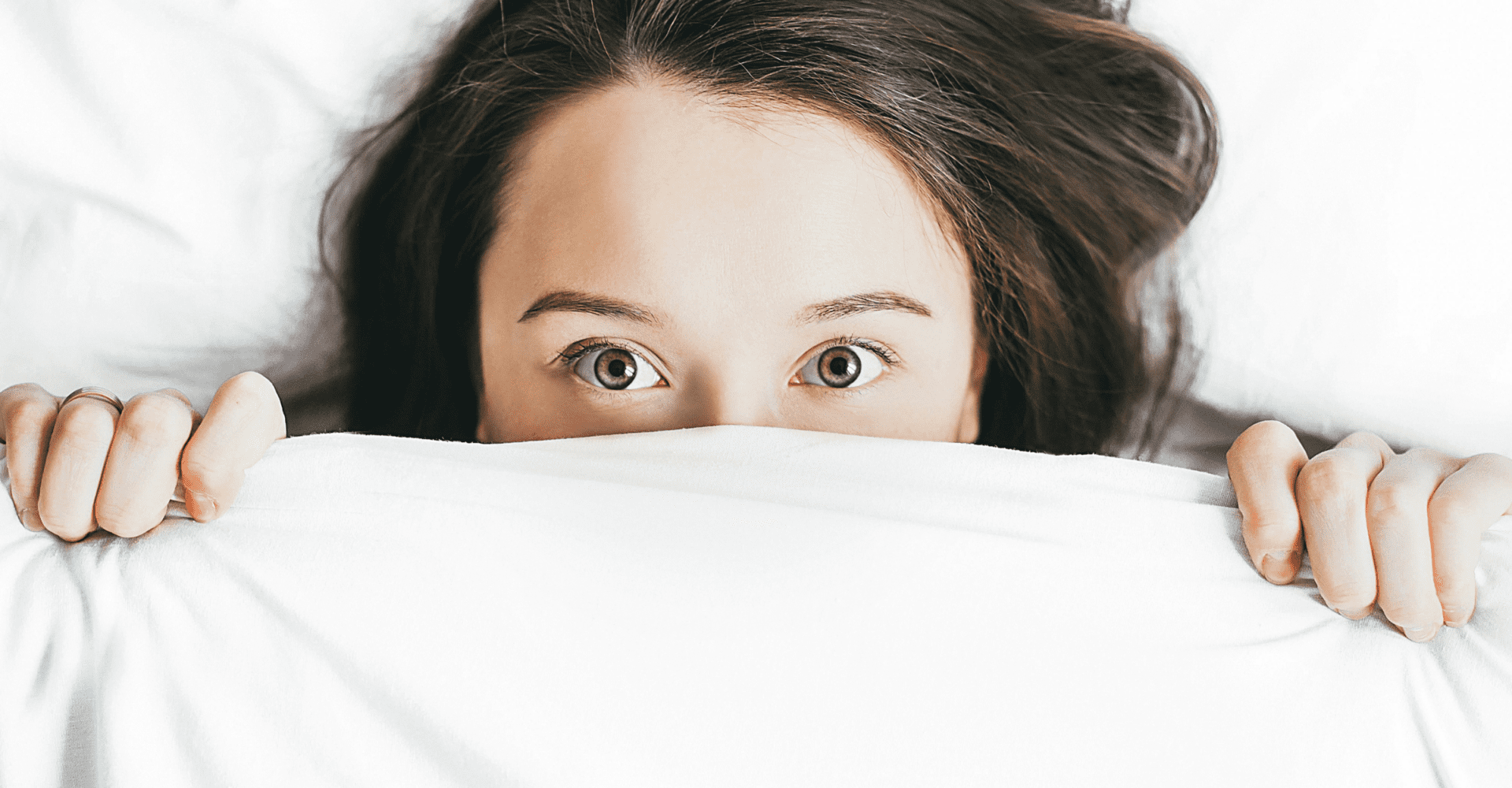 Kobieta z brązowymi włosami i szeroko otwartymi oczami, leżąc w łóżku, zasłania pół swojej twarzy białą kołdrą