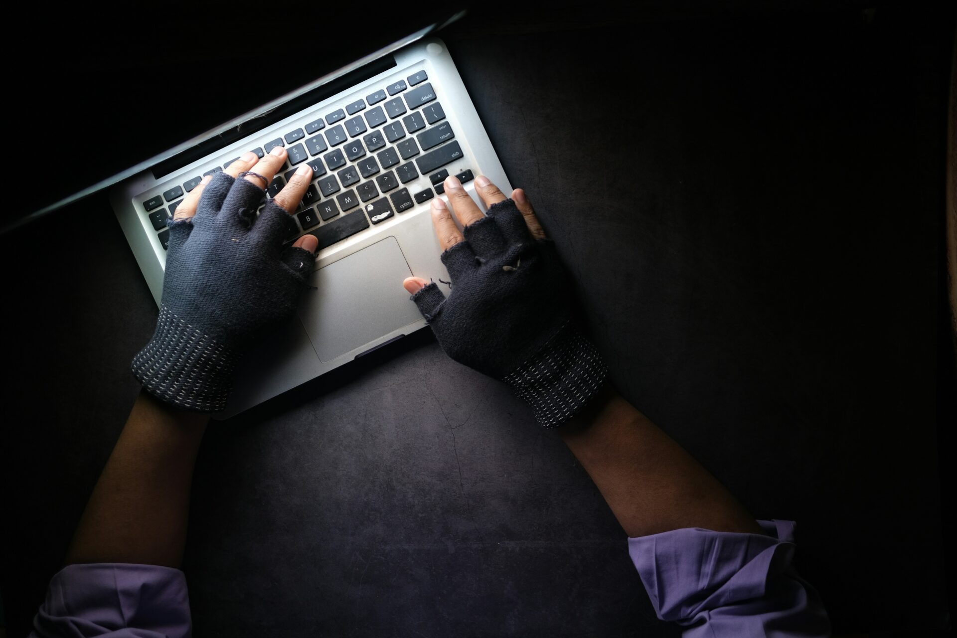 ręce w czarnych rękawiczkach piszą coś na laptopie w ciemnym pokoju
