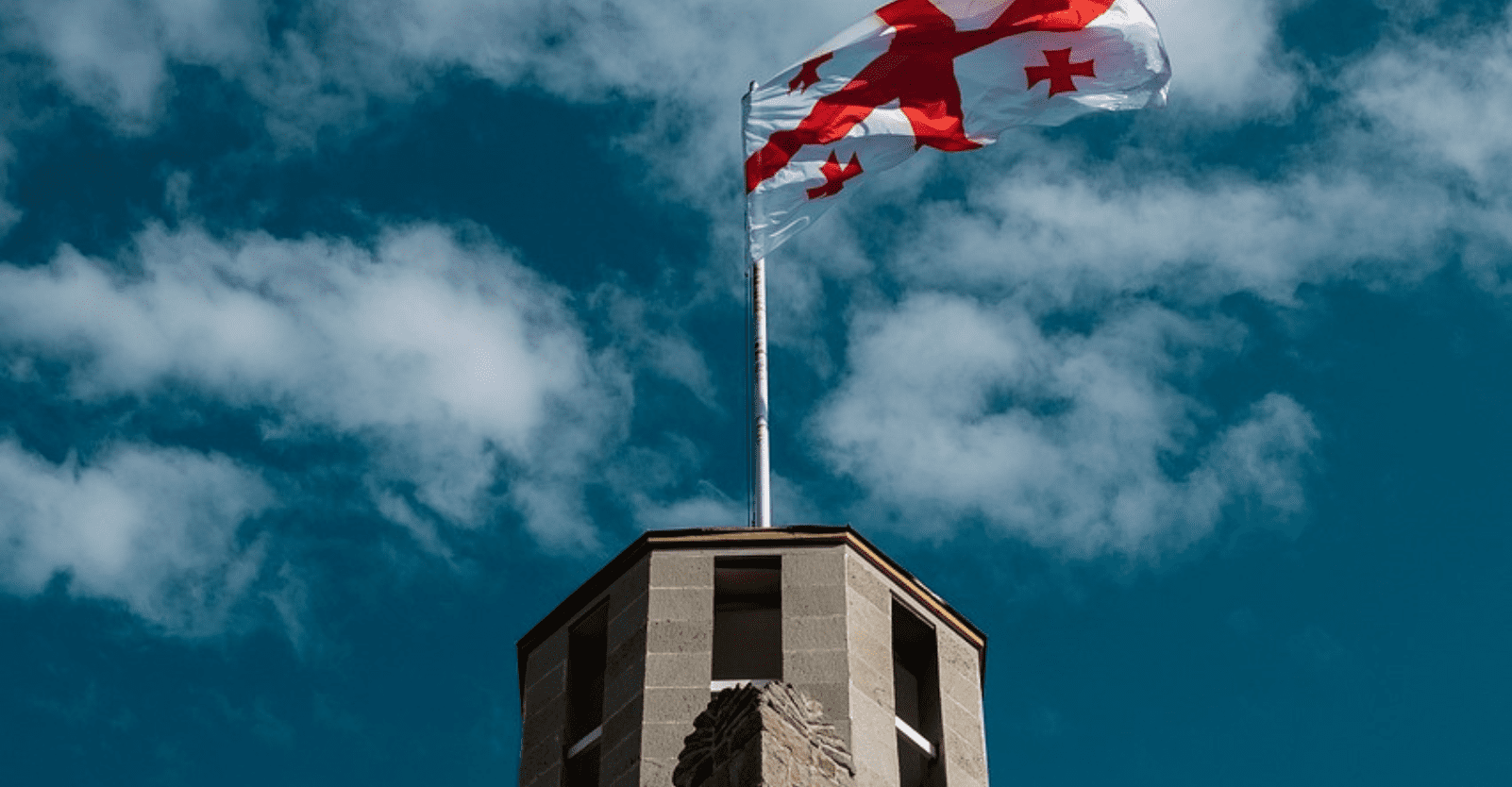 Niebieskie niebo z białymi chmurami. Na środku widnieje budowla z flagą Gruzji (biała flaga z czerwonym krzyżem i małymi czerwonymi krzyżami w każdym rogu).