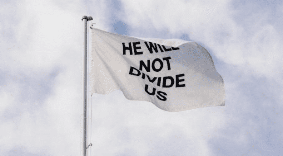 Zdjęcie białej flagi z napisem „We will not divide us”, która powiewa na wietrze. W tle widać zachmurzone niebo.