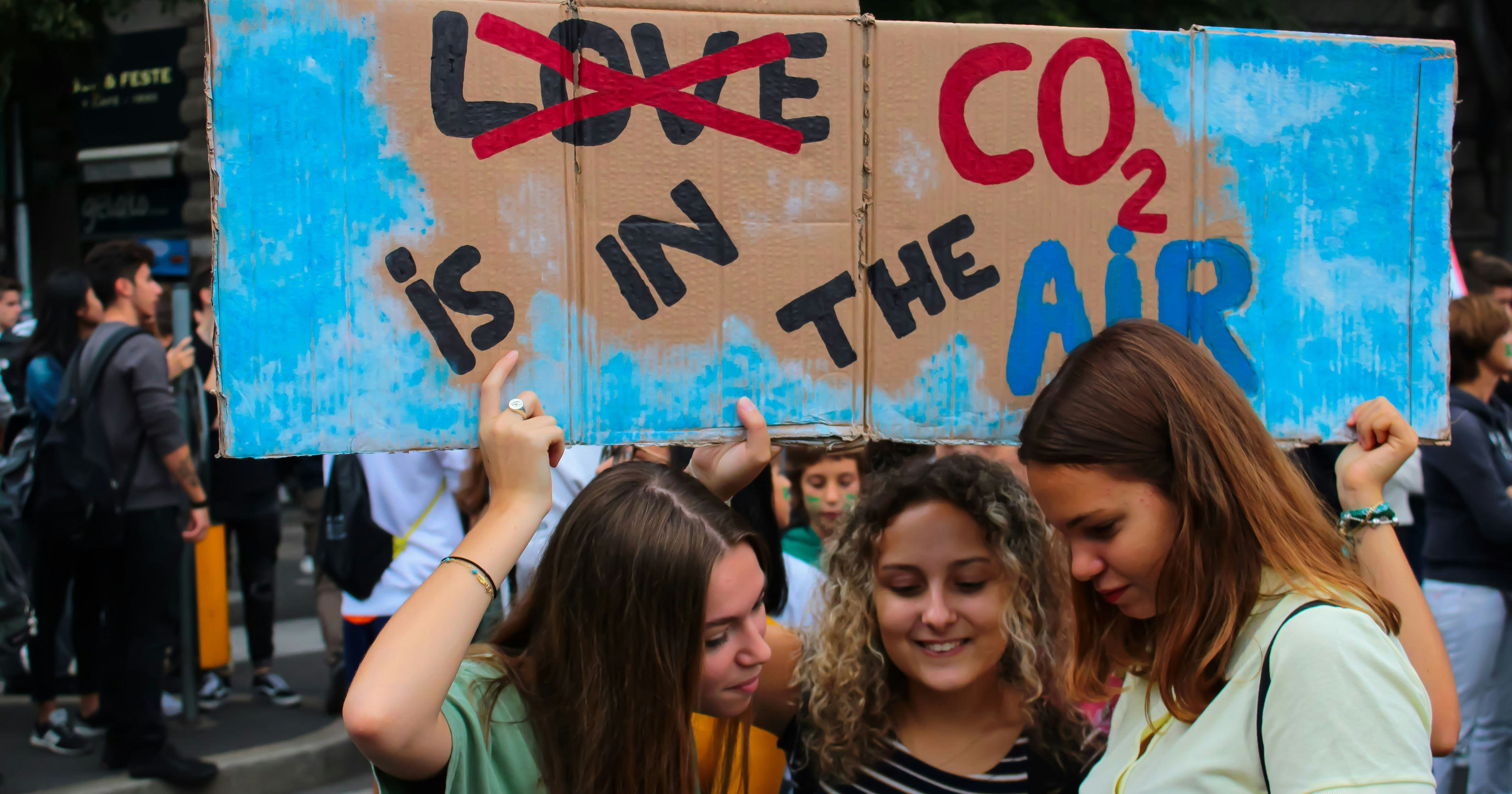 Trzy młode dziewczyny trzymają w ręku karton z przekreślonym napisem "love" i nieprzekreślonym "CO2 is in the air"