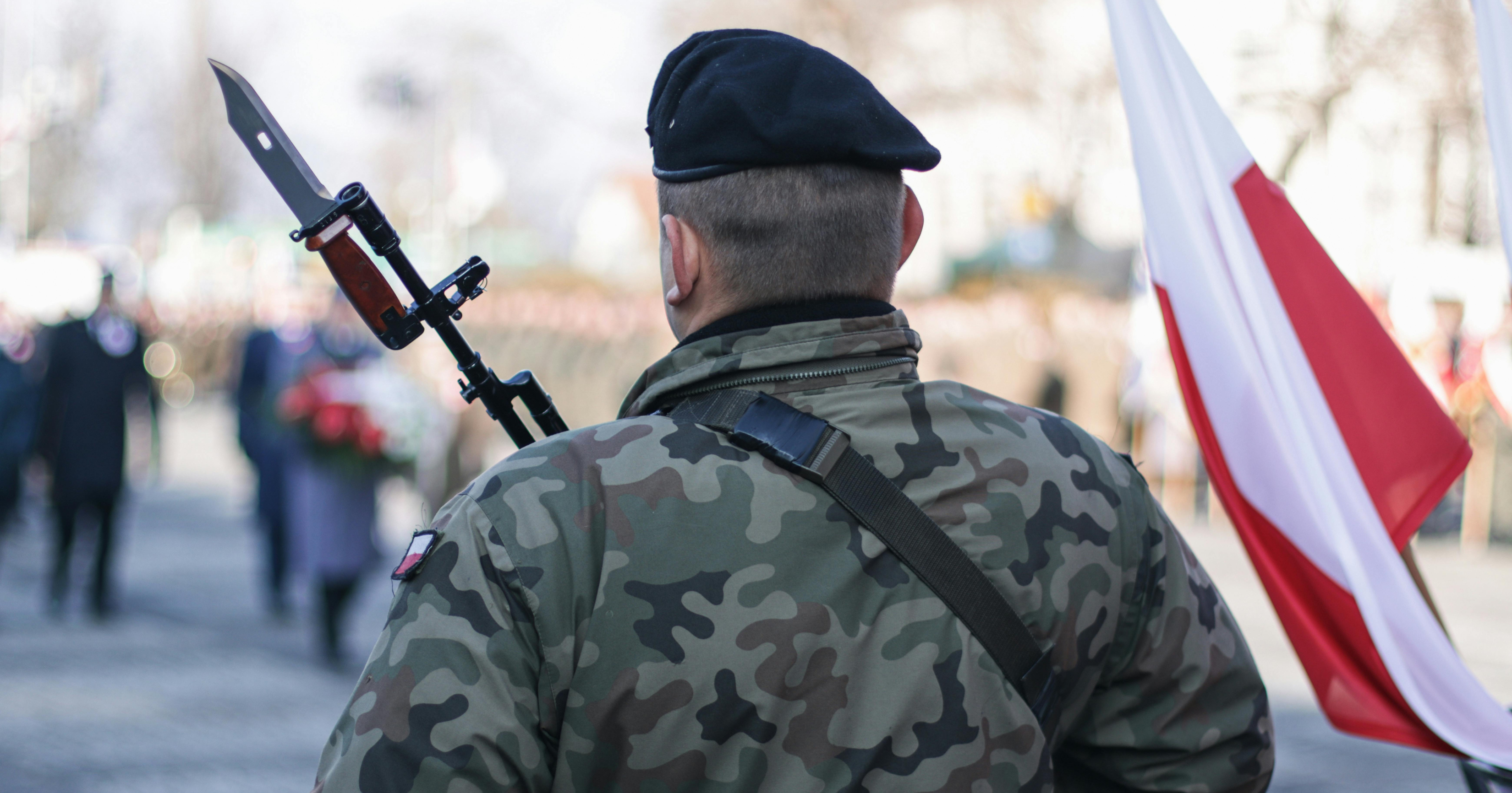 Żołnierz z bronią stoi obok biało-czerwonej flagi Polski.