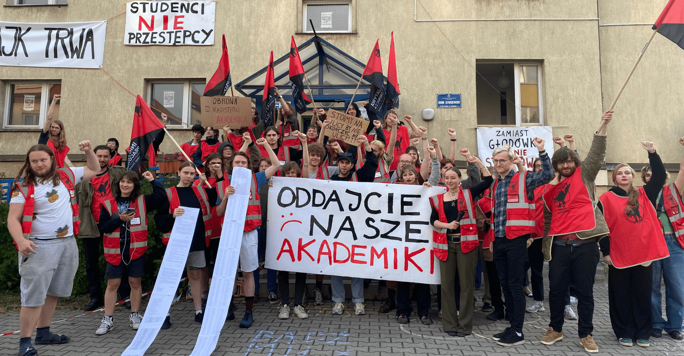 Zdjęcie przedstawiające osoby studiujące, które protestują pod Domem Studenckim Kamionka w Krakowie. Mają na sobie czerwone kamizelki. Kilka osób w rękach trzyma flagę Ogólnopolskiego Związku Zawodowego „Inicjatywa Pracownicza”. Dwie osoby trzymają w rękach długie kartki, które opadają na ziemię. W centrum kilka osób trzyma plakat z napisem: „Oddajcie nasze akademiki”.