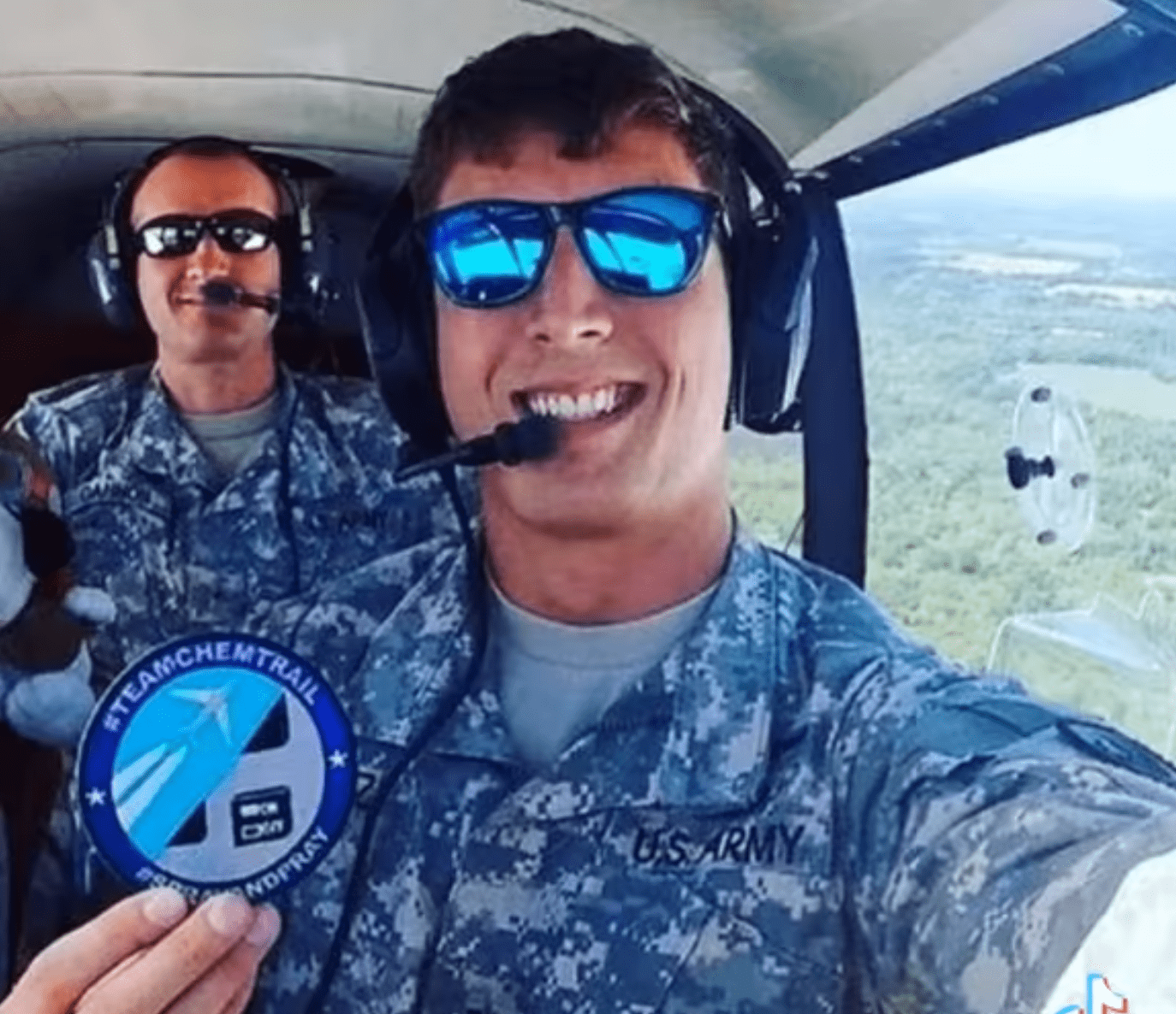 Zdjęcie, na którym widać dwóch mężczyzn w samolocie. Mają na sobie mundury armii amerykańskiej, okulary i zestaw słuchawkowy dla pilotów. Mężczyzna na pierwszym planie trzyma plakietkę z napisem „#TEAMCHEMTRAIL #SPRAYANDPRAY”. 