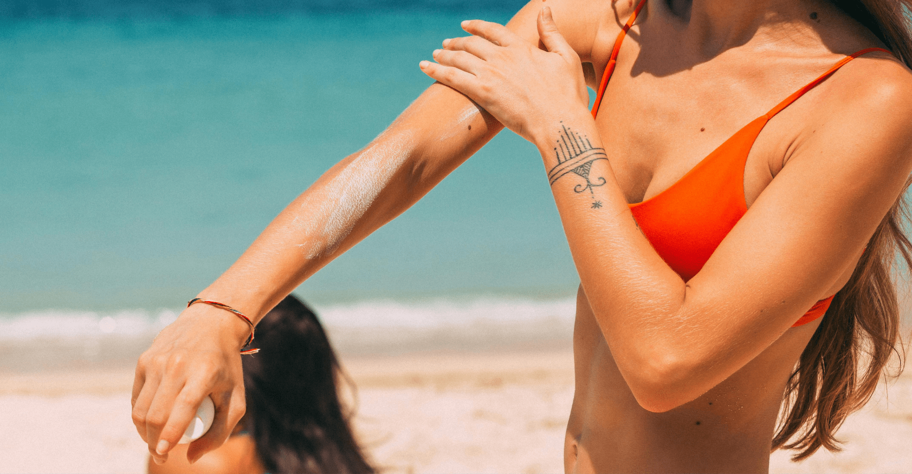 Kobieta ubrana w czerwone bikini smaruje sobie ramię kremem z filtrem. Znajduje się na plaży — w tle widać wodę i piasek oraz inną osobę z długimi włosami.