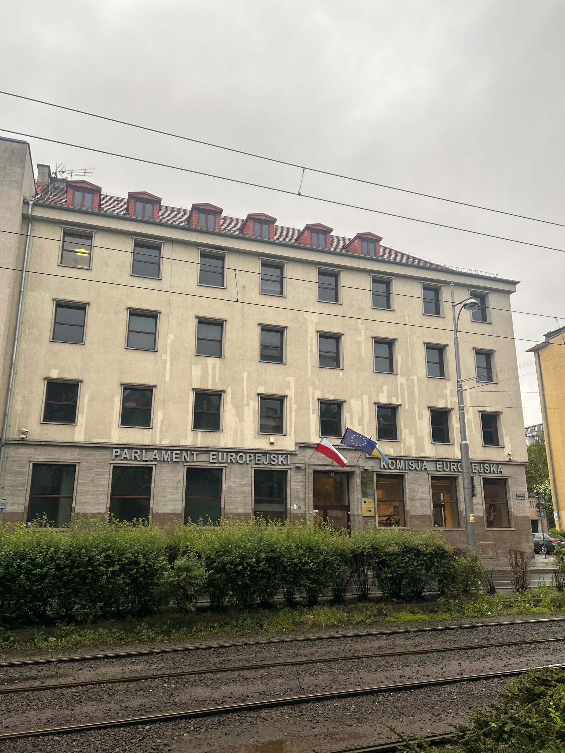 Zdjęcie frontowej części budynku wrocławskiego biura Parlamentu Europejskiego. Nad wejściem widać flagę Polski, Unii Europejskiej oraz Ukrainy.