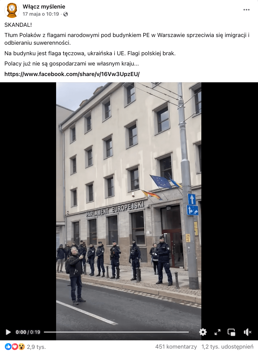 Zrzut ekranu przedstawiający post z Facebooka. Do wpisu dołączono nagranie. W kadrze widocznym na screenie, widać wejście do biura kontaktowego Parlamentu Europejskiego. Nad wejściem powiewają trzy flagi - progresywna LGBTQ+, Unii Europejskiej oraz Ukrainy. Przed gmachem w rzędzie stoją policjanci.