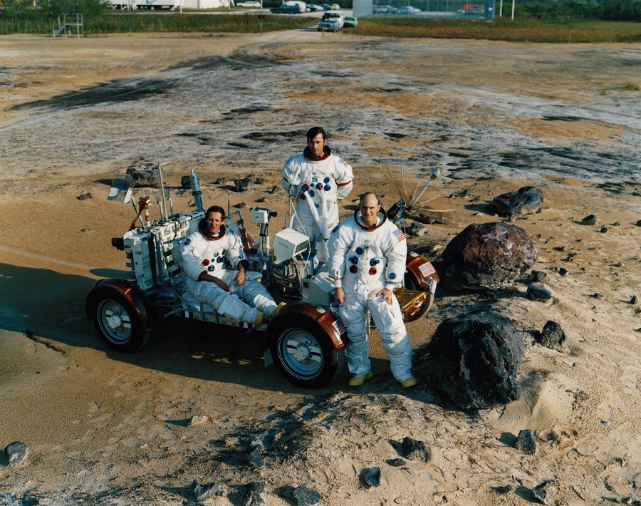 Kolorowe zdjęcie przedstawiające trzech astronautów misji Apollo 16 w trakcie ćwiczeń, stojących obok łazika kosmicznego ze zdjętymi hełmami. W górze kadru widać samochody i fragment parkingu.