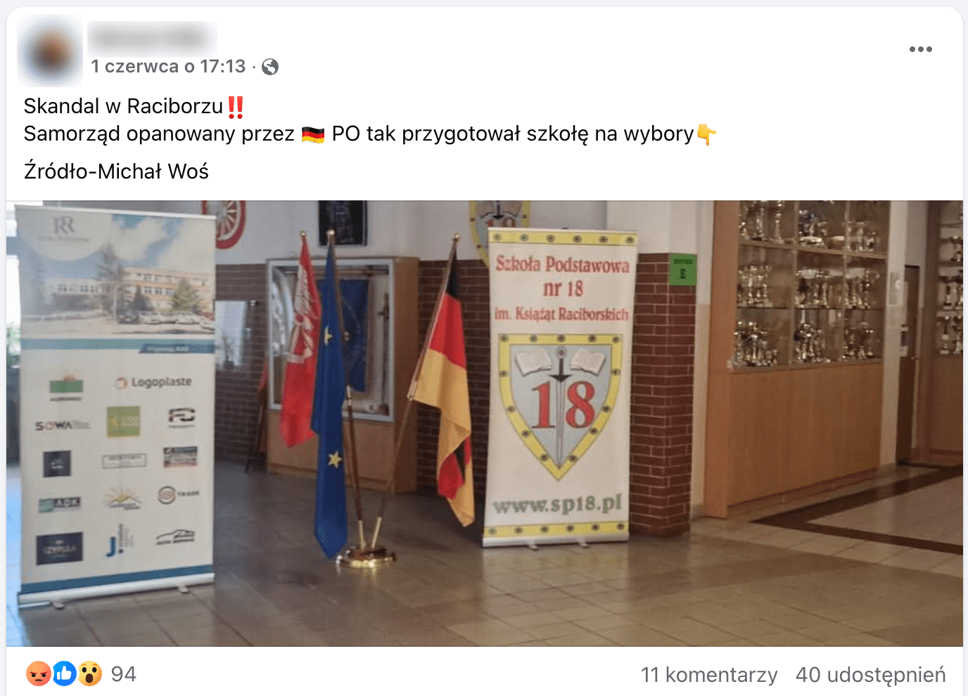 Zrzut ekranu posta na Facebooku. Załączone zdjęcie przedstawia korytarz szkolny. Pod ścianą stoi gablota z pucharami. Na środku ustawiono dwa banery oraz trzy flagi: Unii Europejskiej, Niemiec. Trzecia flaga ma czerwoną barwę oraz biały emblemat, trudno określić, co przedstawia.