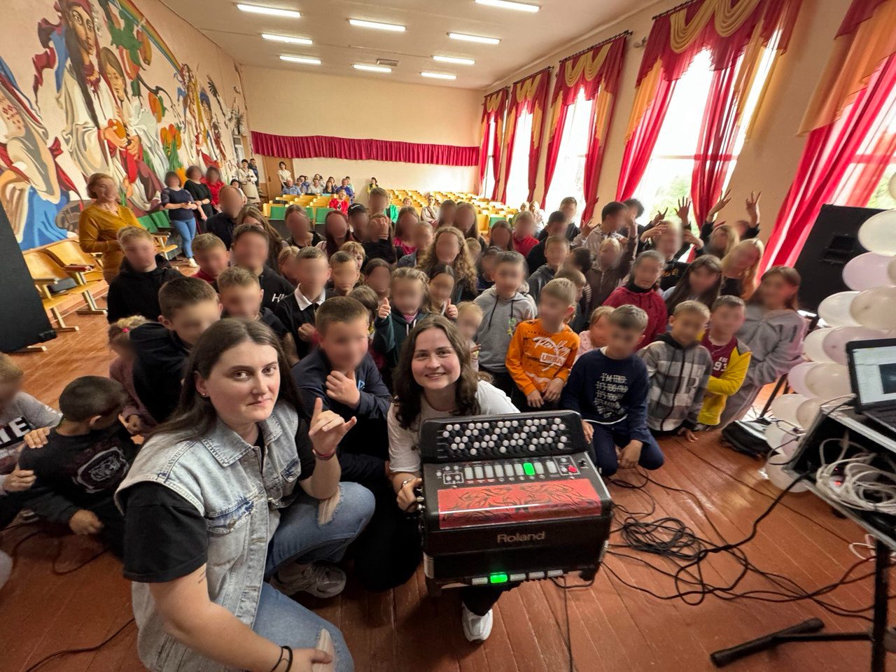 Жінка з акордеоном та інша жінка позують для фотографії в кімнаті, повній дітей, з картиною на стіні та повітряними кульками.